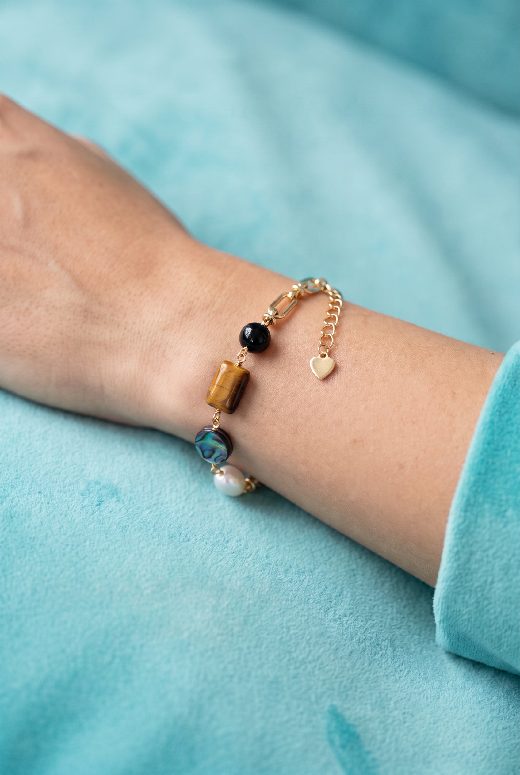 Gold chain bracelet with tiger's eye, obsidian, pearl, and iridescent abalone stones on cream background.