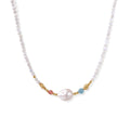 Delicate white beaded necklace with baroque pearl center, gold accents, and coral and aqua gemstones on cream background.