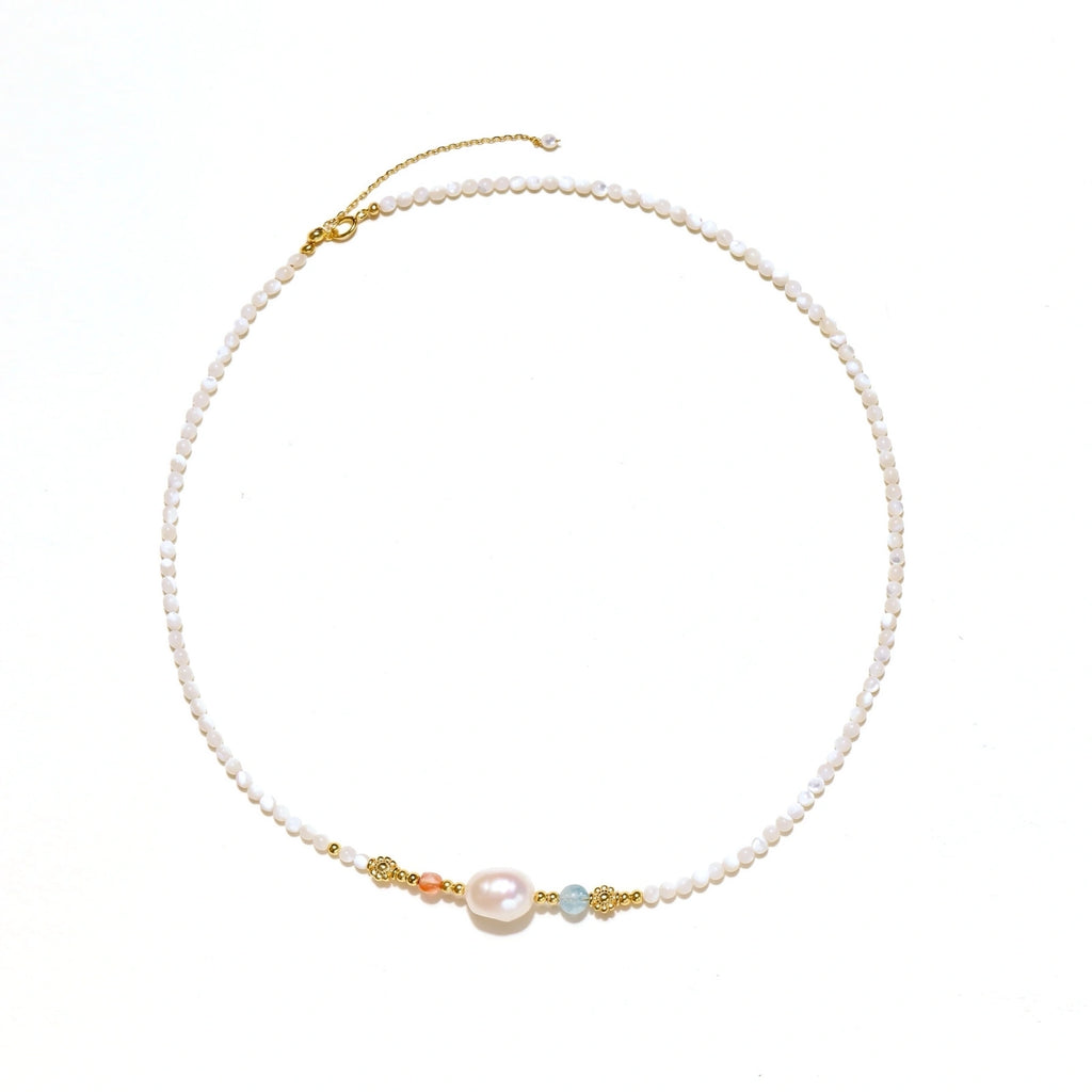 Delicate white beaded necklace with baroque pearl center, gold accents, and coral and aqua gemstones on cream background.