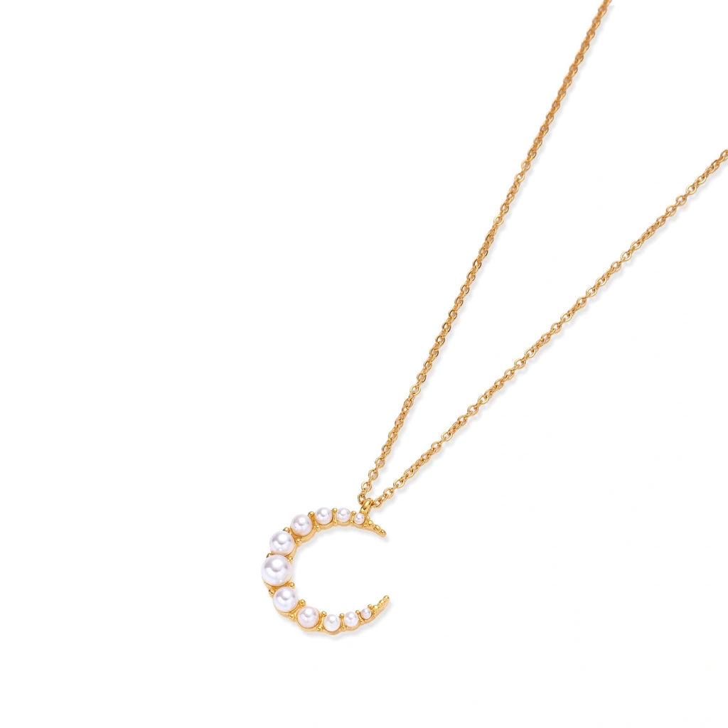 Gold crescent moon pendant with graduated white pearls on delicate chain against cream background