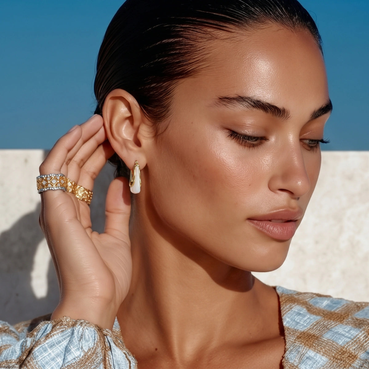 Woman wearing gold earrings and rings against a clear blue sky.