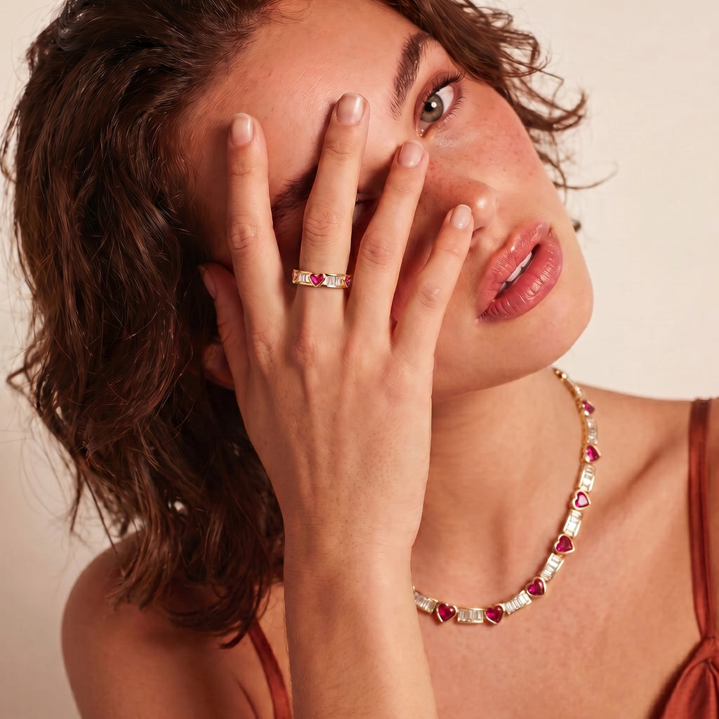 Woman wearing a gold necklace with red gemstones and a ring, against a neutral background