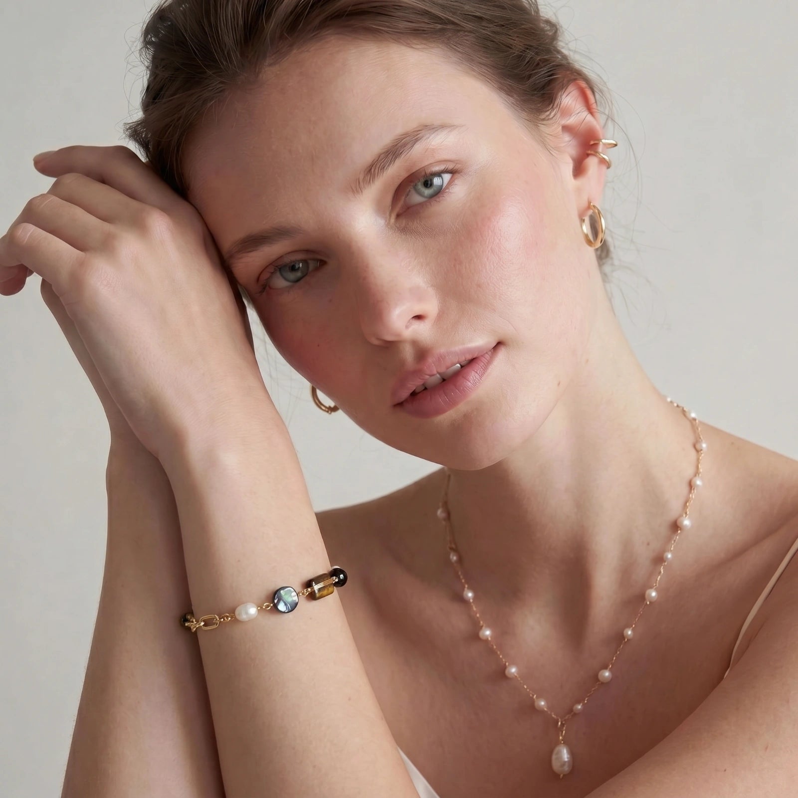 A close-up lifestyle shot of a woman wearing a delicate gold and pearl bracelet on her wrist, along with a matching pearl necklace and gold hoop earrings, set against a soft, light gray background.