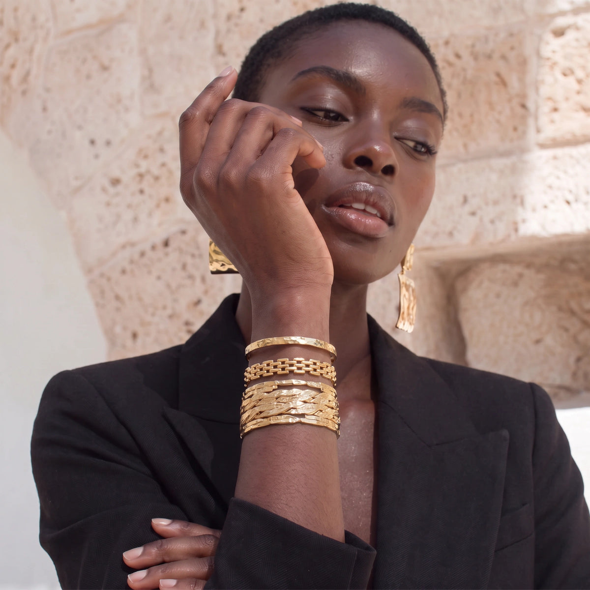 A close-up lifestyle shot of a model wearing a stack of textured gold bangles on her wrist, along with matching gold earrings, set against a light beige stone wall background.