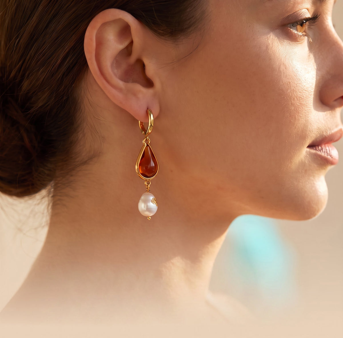 A close-up side profile of a woman wearing a gold drop earring with a teardrop-shaped amber-red stone and a dangling white pearl.